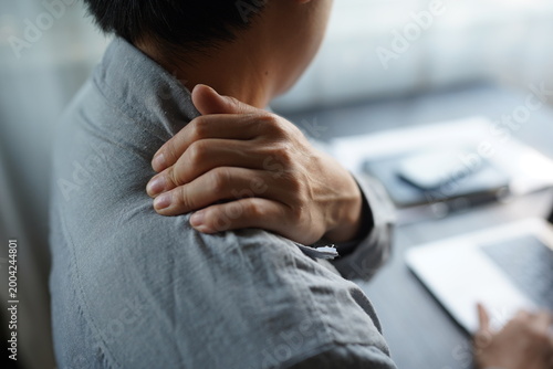 Young man suffering from shoulder and back pain while working on laptop in office.