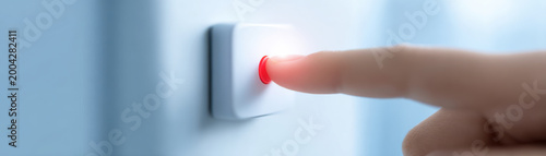 Finger pressing red illuminated button on white wall switch, close up of hand activating control with soft light
