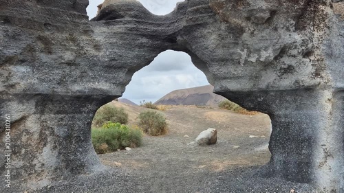 Camera floating through a natural rock arch at Antigua Rofera quarry in Lanzarote, surreal volcanic landscape with carved stone formations, cinematic motion through rugged Canary Islands terrain.