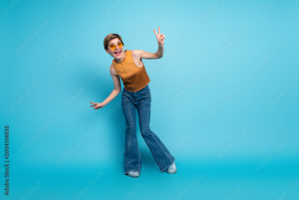 Obraz premium Young stylish woman in orange top and jeans posing with peace sign against blue background