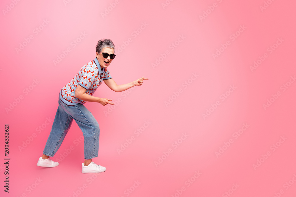 Obraz premium Happy stylish senior woman in sunglasses pointing and laughing against a pink background, casual retro outfit and confident playful pose