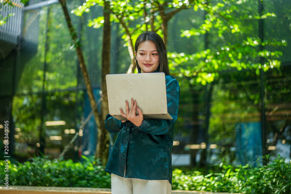 Naklejka premium Professional young Asian woman using a laptop in a lush outdoor park with a modern glass building background. Ideal for remote work, technology, education, and digital nomad lifestyle themes.