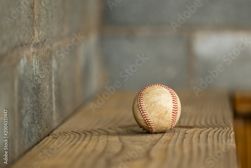 Fotografía Worn baseball sits on a wooden dugout bench in close-up, a weathered cinderblock wall filling the left side of the frame in a textural baseball detail image