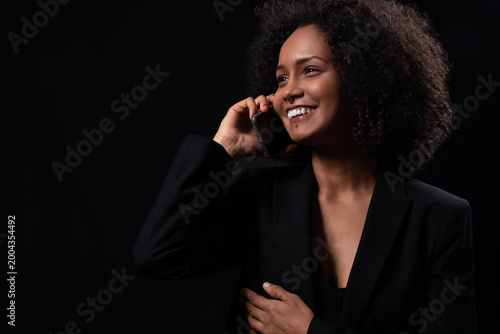 Young woman with afro hair smiling while having a phone conversation on black background