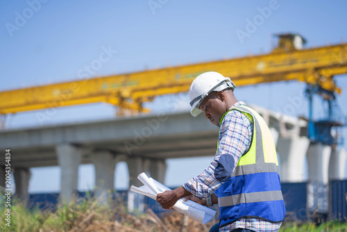 African American civil engineer holding blueprints at a bridge construction site, a professional black foreman in a hard hat and safety vest at an infrastructure project.