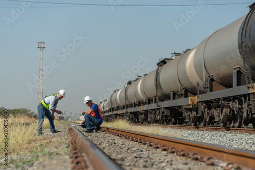 Professional engineers inspecting railway tracks near freight train tankers, railway technicians in hard hats and safety vests checking track alignment.