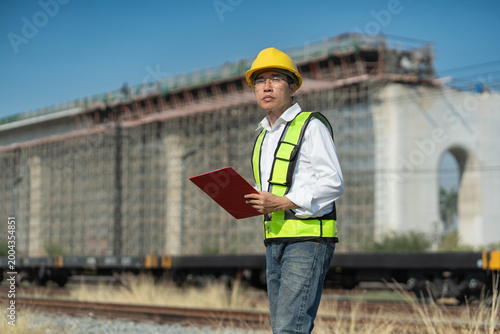Asian civil engineer holding a clipboard at a large bridge construction site, professional foreman in a yellow hard hat and safety vest supervising the project.