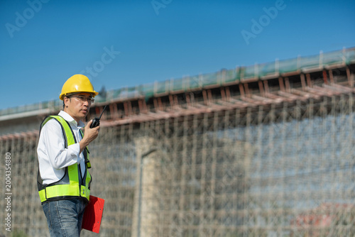 An Asian civil engineer using a walkie-talkie at a large bridge construction site, a professional foreman in safety gear supervising an infrastructure project with a radio.