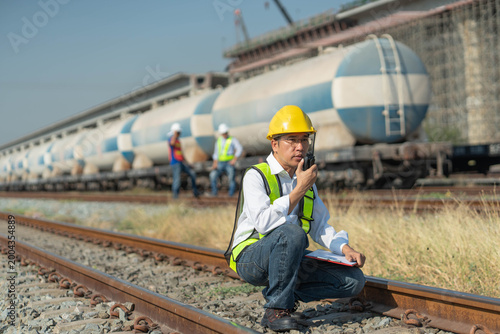A railway engineer using a walkie-talkie while inspecting tracks at a rail yard, a professional asian technician in a safety vest monitoring freight train logistics.