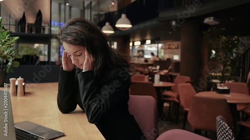Tired young businesswoman working online and using laptop in cafe. Keeping hands on head, massaging of temples. Headache, migraine. Real time