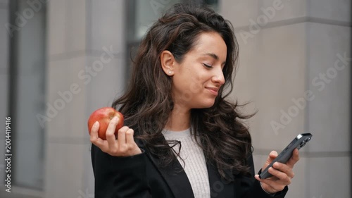 Businesswoman biting an apple while holding smartphone and looking at the screen. Young lady standing outdoor and using mobile phone. Lifestyle, food concept. Real time