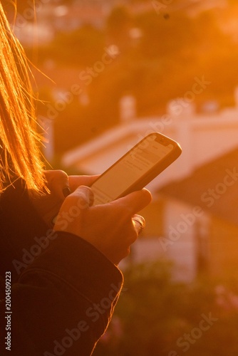 Closeup of a person holding a smartphone outdoors at sunset, with warm golden light, blurred houses in the background, and a casual relaxed atmosphere.