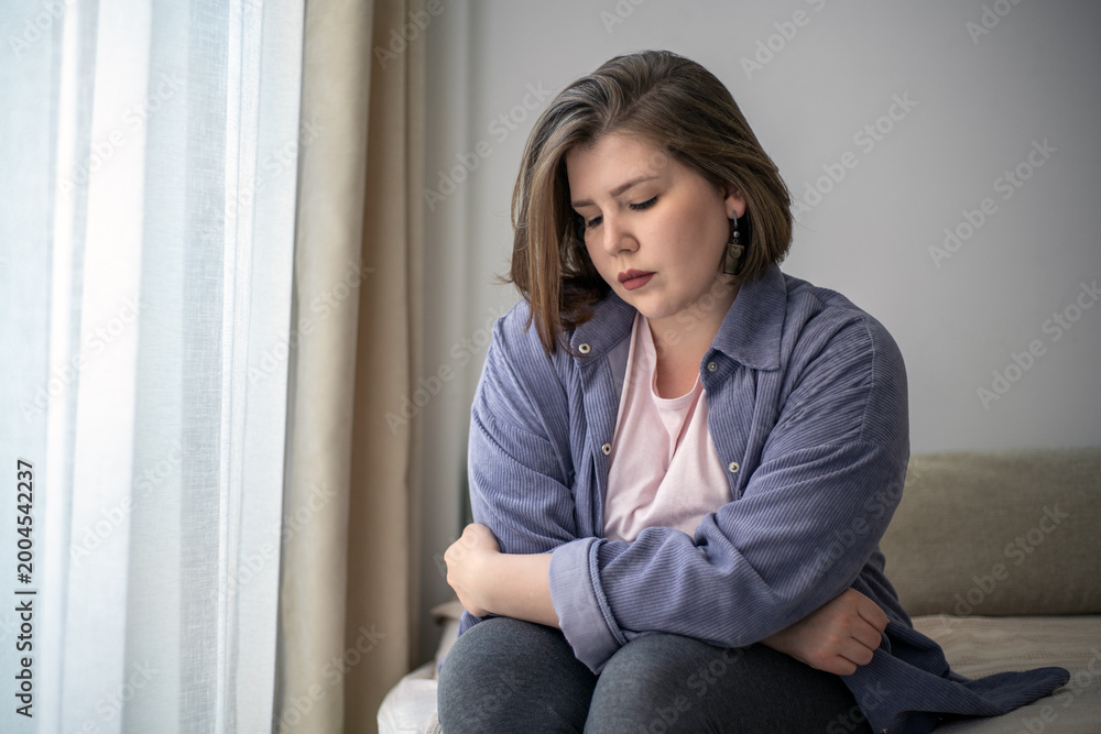 Naklejka premium Lonely plus size woman sitting on bed by window, emotional struggle, anxiety, depression concept, mental health awareness, body positivity.