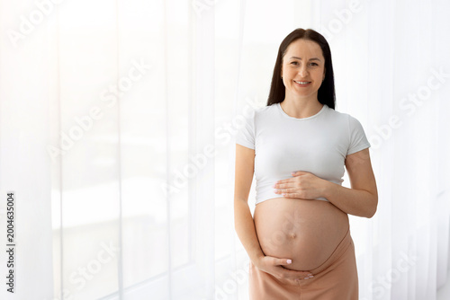 Pregnant young caucasian woman smiling while holding belly, standing against light background, representing happiness, wellness and positive motherhood lifestyle concept Free Download Pregnant young caucasian woman smiling while holding belly, standing against light background, representing happiness, wellness and positive motherhood lifestyle concept - GFXLoad
