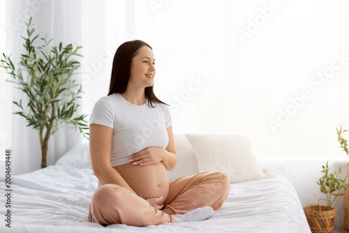 Εκτύπωση καμβά Happy pregnant woman sitting cross legged on bed with bare belly, smiling and lo