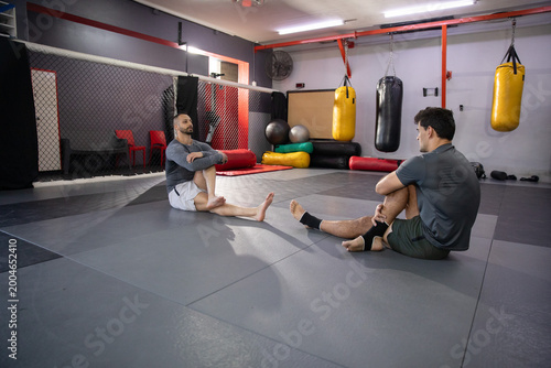 Diverse male partners sitting and stretching on gray mats at martial arts gym with punching bags