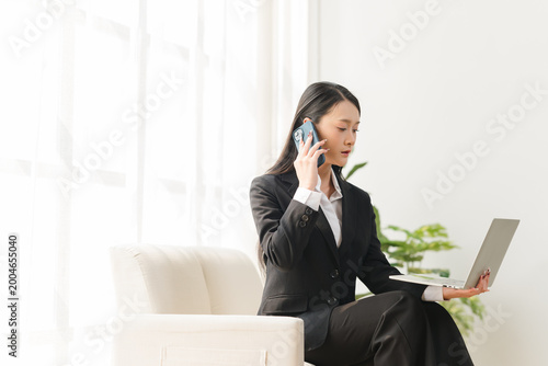 Foto Businesswoman on a Call: A focused businesswoman in a sleek suit manages multiple tasks with finesse, engaging in a phone conversation while working on a laptop in a bright and airy room
