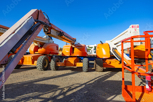 Row of orange telescopic boom lifts parked on a construction site under a clear blue sky in bright daylight