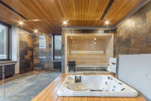 Luxury home spa interior with wooden sauna, jacuzzi hot tub and stone walls.Modern wellness area features glass sauna, white jacuzzi, and walk in shower. Natural wood and stone materials create relax