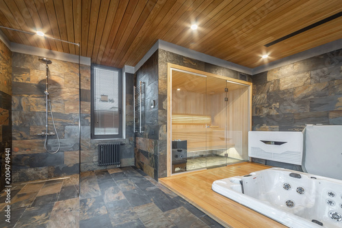 Foto A glass-enclosed sauna with light wood interior paneling and built-in lighting sits behind the hot tub