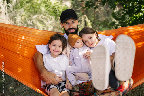 Family enjoys relaxed time together in bright orange hammock under sunny trees in peaceful outdoor setting