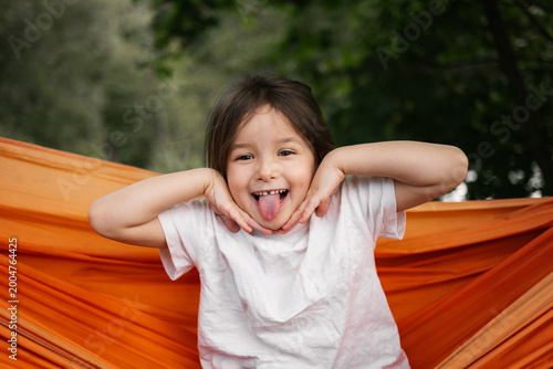 Young girl laughs with tongue out in bright orange hammock surrounded by green trees outdoors