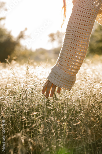 Hand in soft knitted sleeve brushes tall grasses as golden sunlight warms the field and peaceful moment unfolds