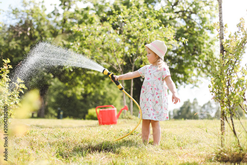 A little cute baby girl 3-4 years old in a dress watering the plants from a watering can in the garden. Kids having fun gardening on a bright sunny day. Outdoor activity children