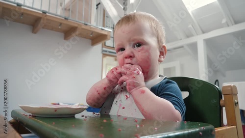 Messy Infant eating cookie at home. Dirty and cute baby with food stains sits happily in high chair and looks at camera, clearly demonstrating the pure joy and inevitable mess that comes with eating
