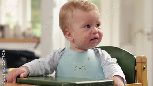 White baby in hight chair reacting with puzzled and fussy expressions, wearing pastel bib, sitting at wooden table in bright kitchen, natural sunlight from window