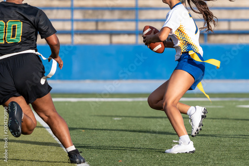 Female flag football ball carrier running past a defender during a competitive outdoor game, with yellow flags and football visible in a dynamic sports action scene.