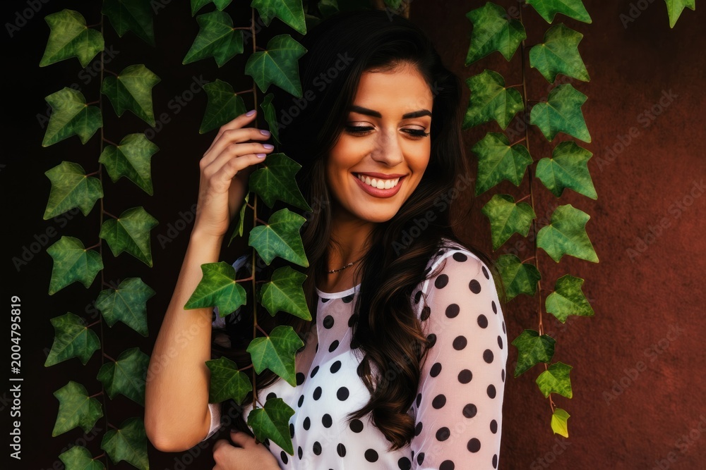 Fototapeta premium Young Latina woman smiles behind green ivy. She wears a white polka dot sheer shirt. Her dark hair flows as she touches a plant leaf. Natural beauty outdoor shot.