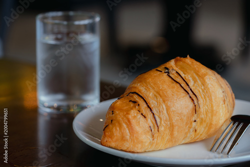 Konstfotografi A golden croissant with chocolate drizzle served on a white plate, accompanied by a fork and a glass of water on a wooden table in a cozy café setting
