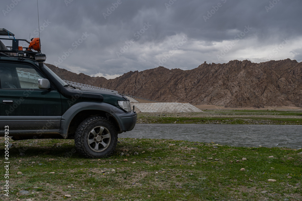 Fototapeta premium Off-road 4x4 vehicle with expedition roof rack parked near a mountain river. Adventure travel and overlanding concept in wild rocky landscape under cloudy sky.