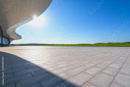 A bright sunny open square paved with stone tiles, featuring a curved modern building facade, wide green lawn under clear blue sky.