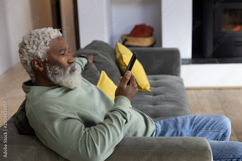 Obraz premium Senior African American man reclining on gray sofa in living room, holding phone near fireplace
