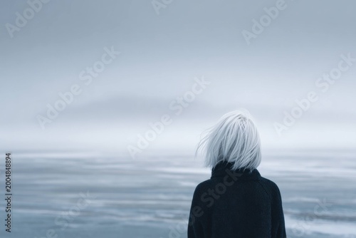 Woman with white hair wearing black coat stands alone facing calm sea under cloudy sky, showing solitude and contemplation in cold weather outdoors.