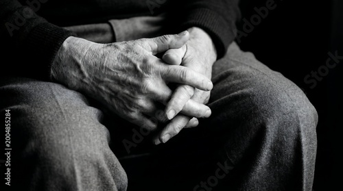 Close-up black and white portrait of an elderly person's hands clasped together on their lap with a somber mood.