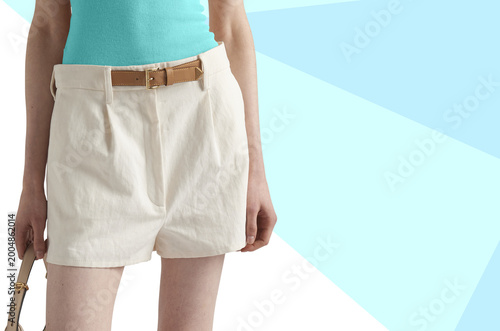  Close-up of a fresh summer outfit featuring white high-waisted shorts paired with a turquoise sleeveless top and a brown leather belt. The model holds a handbag, set against a soft pastel geometric 