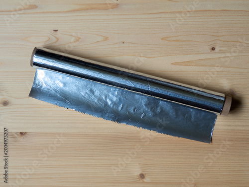 Roll of aluminum foil with a serrated cutting edge partially unrolled on a light wooden surface. Household kitchen item used for food storage, wrapping, and cooking