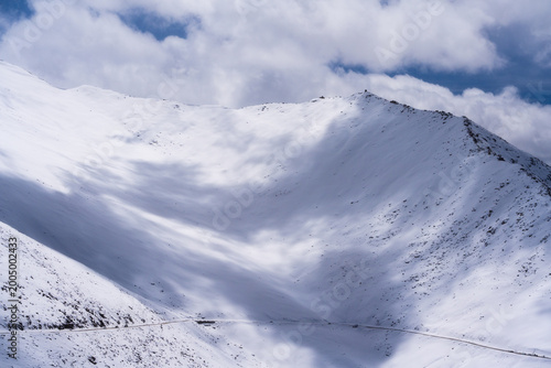 Majestic snow-covered mountain slope in Ladakh, India, with a small winding road at the base. High altitude winter landscape featuring soft light and cloud shadows. Minimalist nature travel background