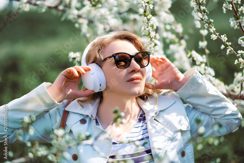 Woman in sunglasses and white headphones listening to music among white blooming trees. Audio enjoyment and nature harmony concept