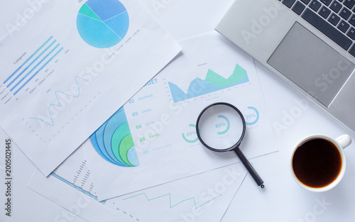 Printed charts and graphs with magnifying glass and laptop corner. Concept of business analytics, market research, and data visualization.