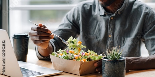 Man eating a fresh salad from a takeout container while working on his laptop at a table with a coffee cup and potted plant