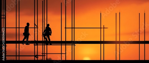 Silhouetted construction workers on scaffolding against a vibrant orange sunset sky, symbolizing hard work and development