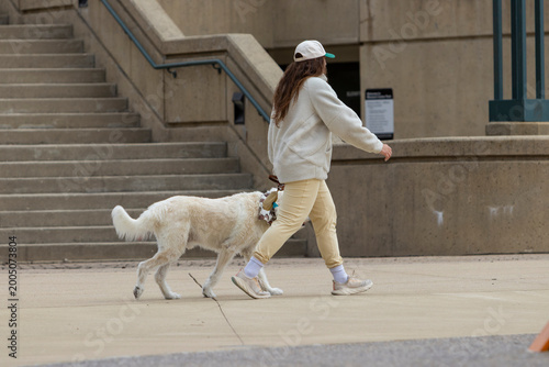 A woman with long dark hair and a tan hoodie and pants, walking a white and tan dog along a concrete sidewalk next to a wall and stairs in the same color scheme.