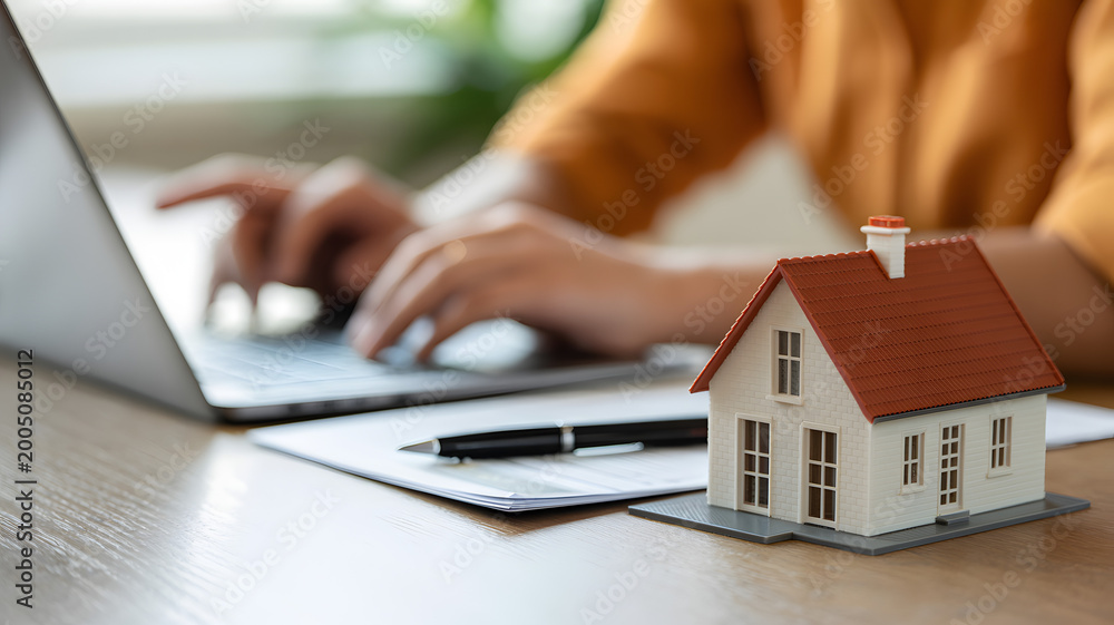 Fototapeta premium Woman typing on laptop with miniature house and pen on desk during remote work