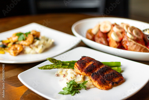 Three white plates on a wooden table featuring an assortment of foods from shrimp and salmon to french toast and bananas, as available in a diner or restaurant.