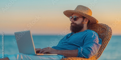 Bearded man wearing hat and sunglasses works on his laptop while sitting in chair on beach during sunset. High quality photo