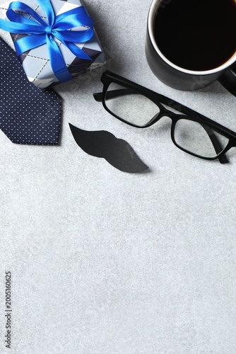Happy Fathers day concept. Gift box, eyeglasses, blue tie, mustaches and cup of coffee on grey backdrop. Top vertical view